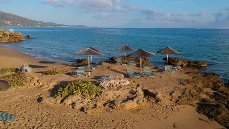 Playa-Rocosa-De-Plaka-Junto-Con-Sillas-Y-Sombrillas-De-Palma-Solar-En-El-Punto-Más-Oriental-De-La-Isla-Griega-De-Zakynthos,-Toma-Aérea-De-Acercamiento-Con-Drones