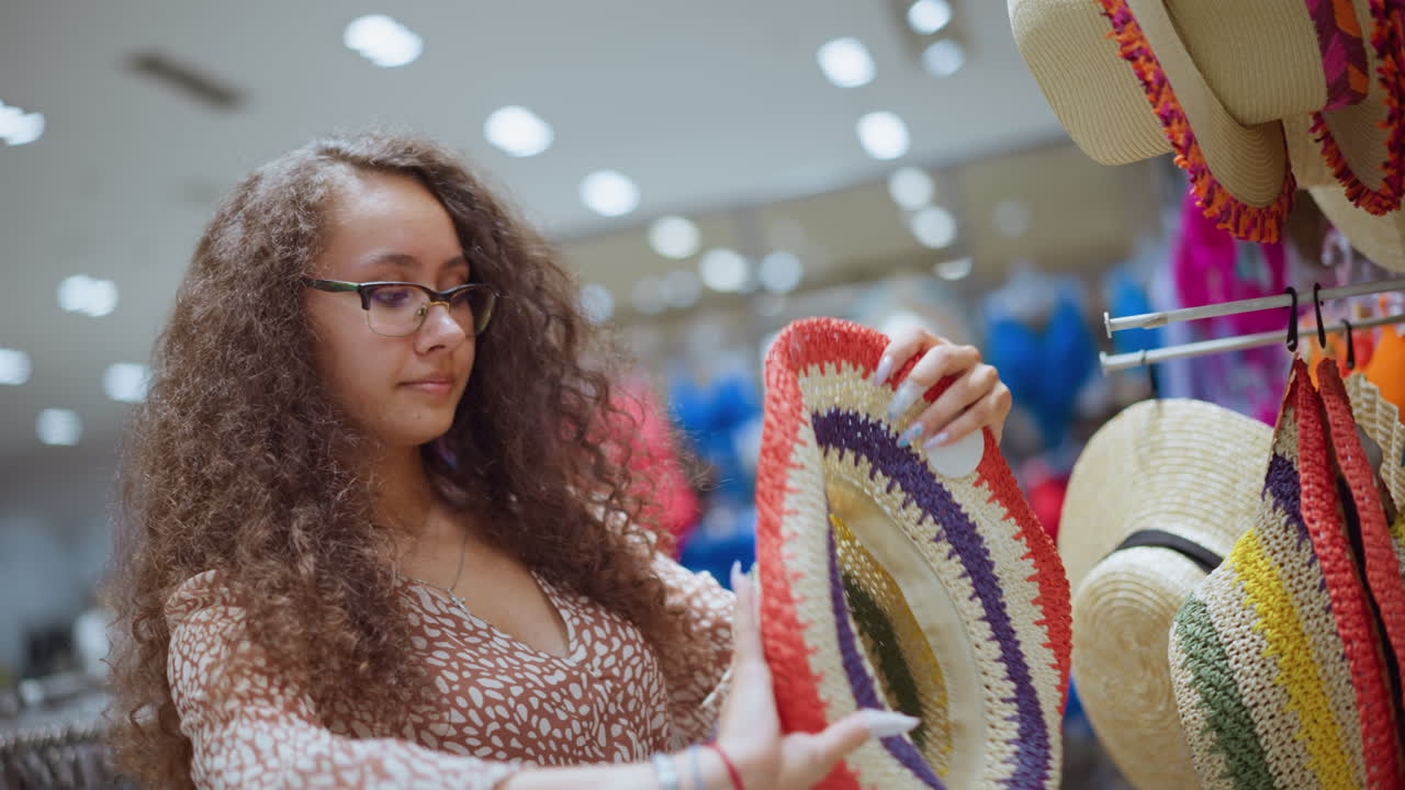 Young woman wearing a handcrafted woven hat with colorful accents poses happily in a brightly lit clothing store, her smile reflects her enjoyment of trying on unique fashion items