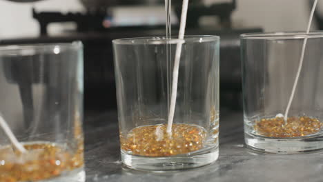 close up of candle wax production process showing person pouring hot melted wax into clear glass cup filled with golden wax beads and wick centered inside