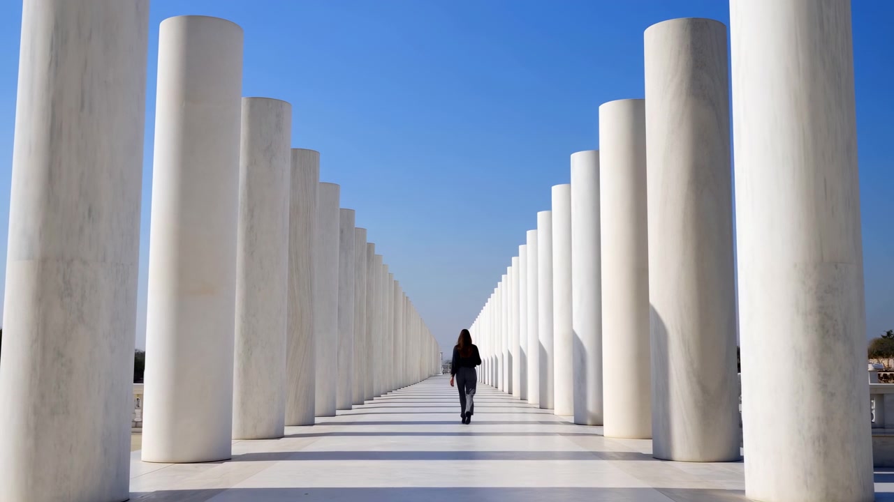 Una donna solitaria cammina lungo il centro di un lungo corridoio simmetrico formato da file di alte colonne bianche. La forte luce solare crea ombre nette sul pavimento luminoso, enfatizzando le linee geometriche, lo spazio aperto e l'architettura monumentale.