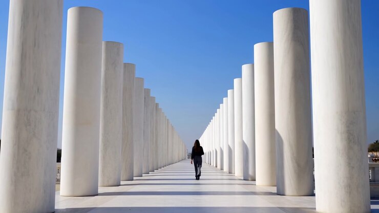 A solitary woman walks along the center of a long symmetrical corridor formed by rows of tall white columns. Strong sunlight creates crisp shadows across the bright pavement, emphasizing the geometric lines, open space, and monumental architecture
