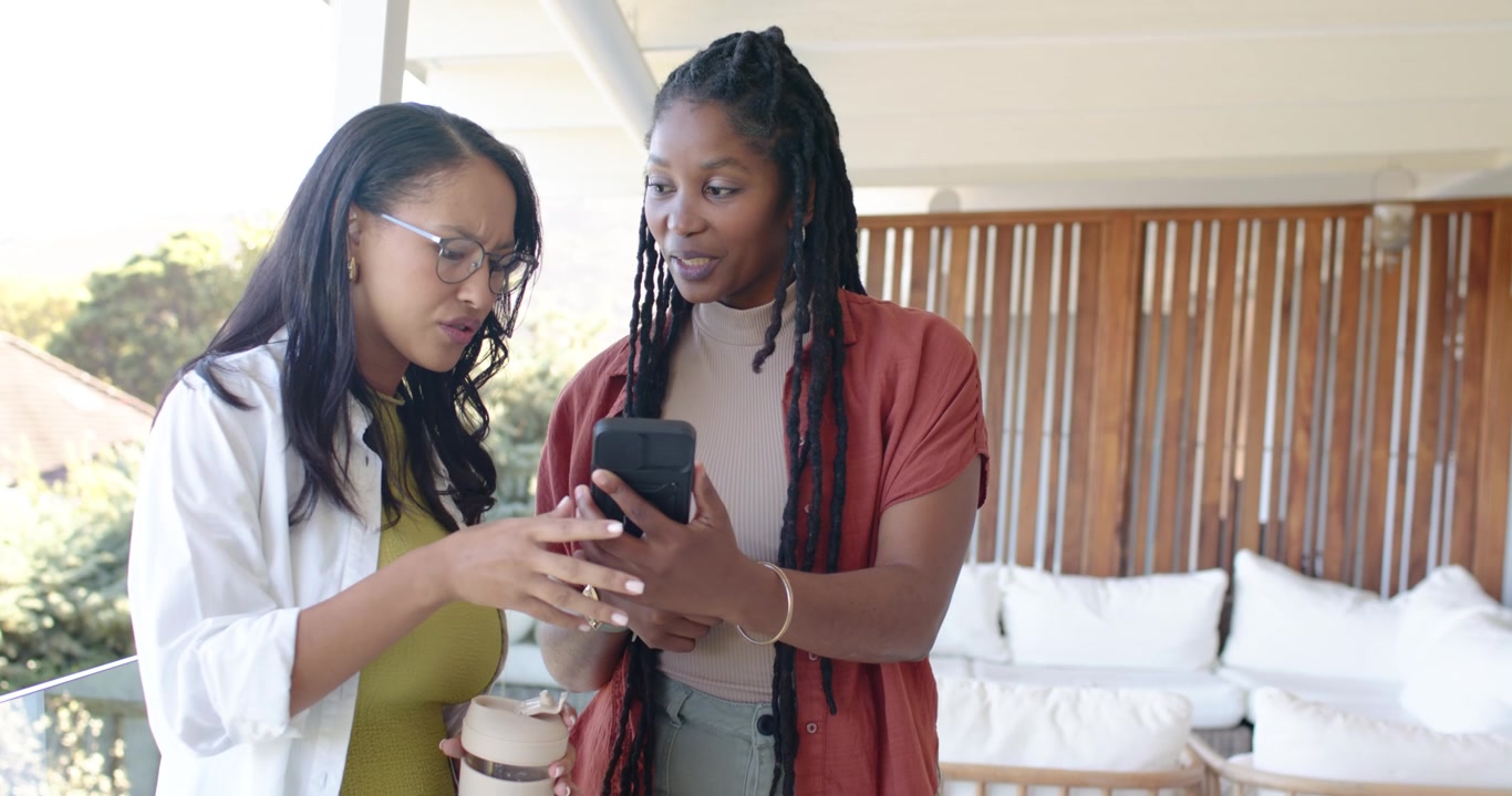 African American women on balcony, right tapping smartphone, left holding coffee cup, sharing plans