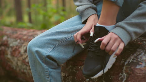 close-up of woman tying shoelaces on black sneakers, wearing gray sweater and light blue jeans, with long white nails, seated on fallen tree in forest