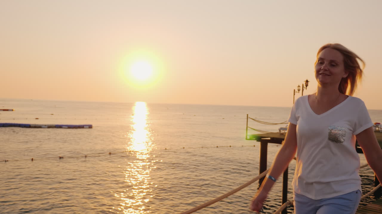 una mujer feliz camina alegremente por el muelle a la orilla del mar con un sombrero en las manos admirando el