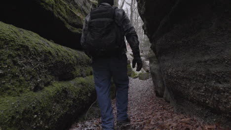 slow-motion-shot-of-a-lone-hiker-wearing-a-backpack-exits-a-cave-through-rocky-moss-covered-walls-and-leaves-covering-the-forest-floor