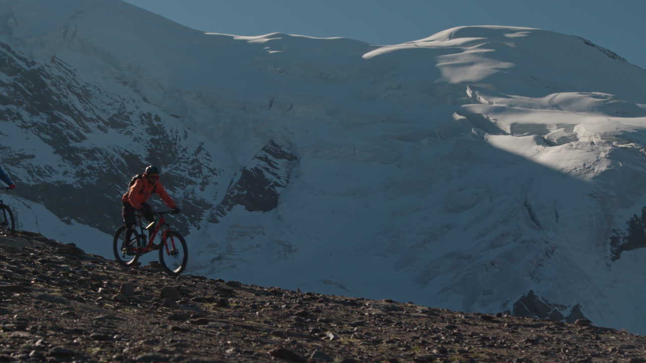Due Mountain Biker Passano Davanti alla Telecamera e Scendono in Discesa con Ampie Viste sulla Valle Alpina a Saas-Fee, Alpi Svizzere. Azione Dinamica sul Sentiero, Scenario Montano Panoramico e Atmosfera Cinematografica di Avventura Estiva