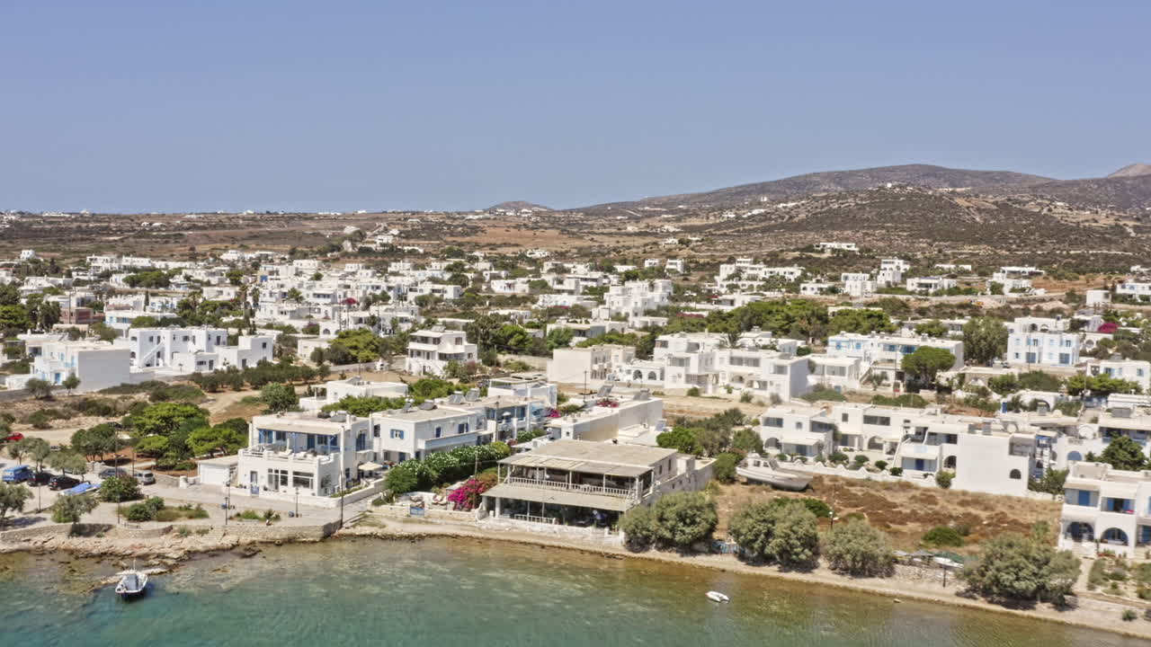 Aliki, Paros, Grecia. Ripresa aerea v2 con zoom indietro che cattura splendidi edifici imbiancati sul lungomare lungo la spiaggia di Paralia con un tranquillo paesaggio marino verso il molo - Settembre 2021.