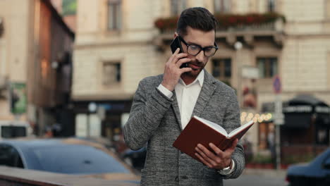 Stylish-Businessman-In-Glasses-Standing-In-The-Street,-Talking-On-The-Phone-And-Looking-At-His-Open-Planner,-Then-Going-Away-1