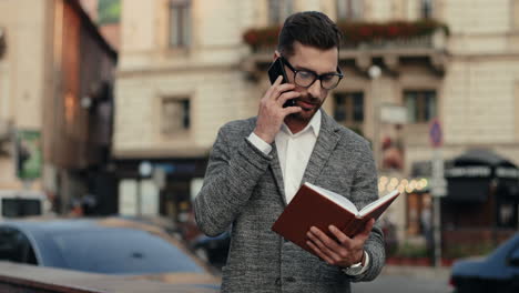 stylish businessman in glasses standing in the street, talking on the phone and looking at his open planner, then going away 1