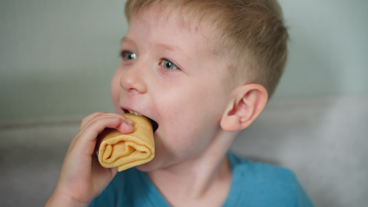 Close up of happy young boy wearing blue shirt smiling joyfully while taking bite of snack indoors carefree spirit and pure happiness of childhood in bright casual home environment