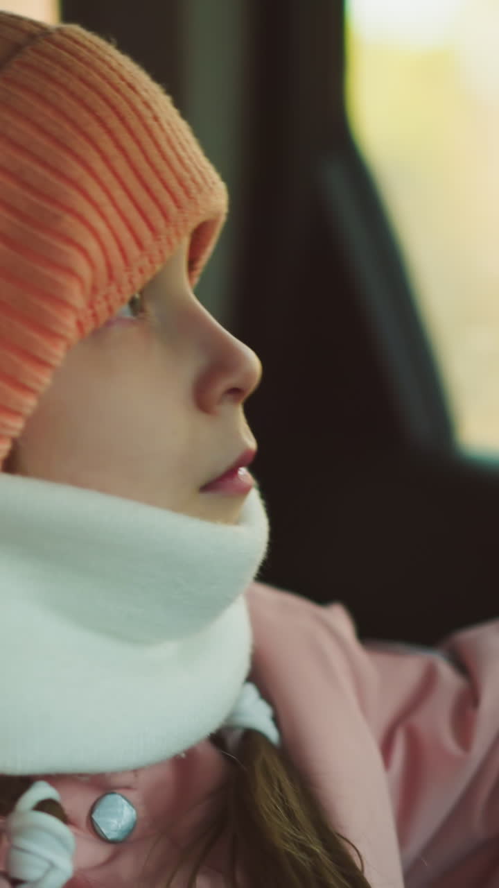 un momento contemplativo como una niña con una gorra rosa, chaqueta y bufanda blanca de invierno se sienta en un coche, colocando suavemente su mano en la ventana
