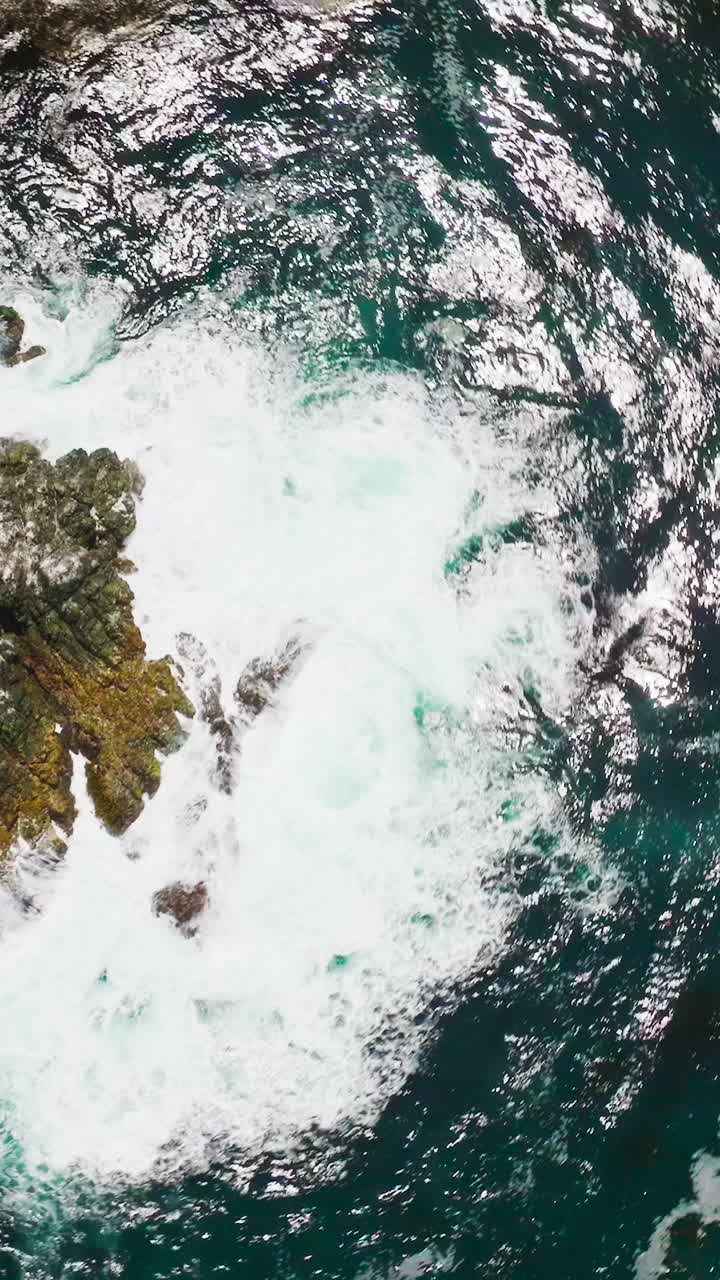 Craggy ragged rock in the blue ocean. White waves splashing by the cliff covered with sea salt. Top view. Vertical video
