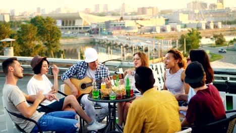 young people are having fun on rooftop playing the guitar, singing, chatting and laughing sitting at table outdoors. joy, music, youth and friendship concept.