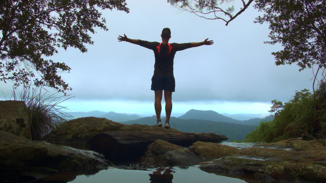 joven parado en la cima de la cascada, disfrutando de la vista a la montaña en el circuito warrie, parque nacional springbrook, australia