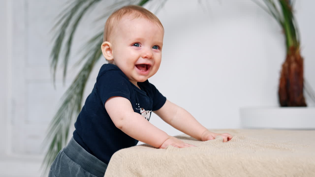 Adorable smiling baby boy stands holding by the sofa. Cute cheerful child waving his hand. Close up.