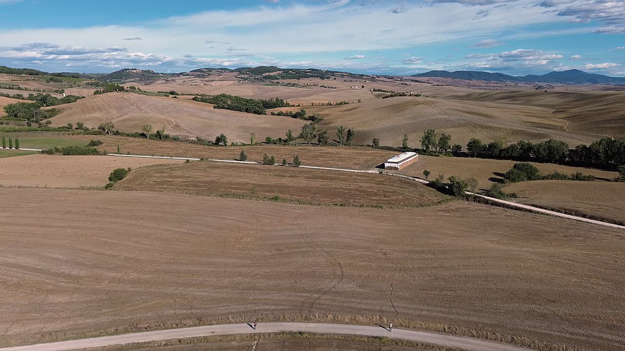 A scenic aerial view of expansive farmlands featuring rolling hills, a curving dirt road, scattered trees, and distant buildings.