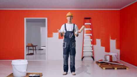 full body of wondering asian man painter wearing safety helmet saying why and standing doubtfully with painting wall in room