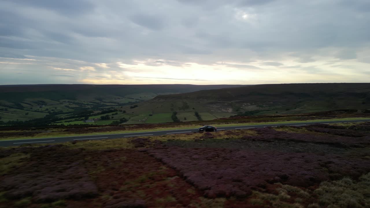 Drone side-tracking a sports car racing along a winding road in North Yorkshire Moors National Park, England during sunset with cloudy sky