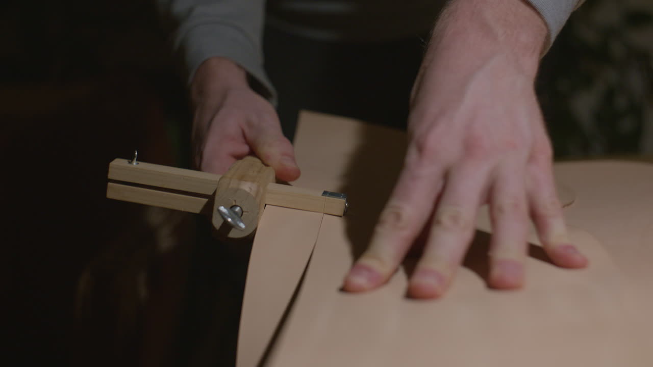 A craftsman carefully guiding a wooden leather edge tool along a leather sheet, applying pressure with his hand for precision.