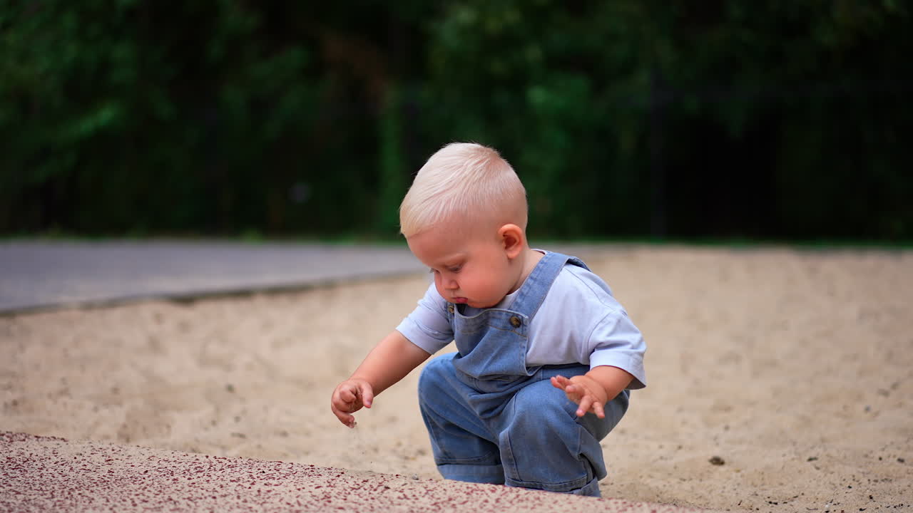 Small baby boy with adorable plump cheeks is on the walk. Lovely child picks some sand and looks at it carefully.