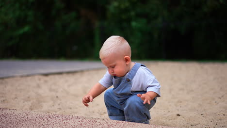 small baby boy with adorable plump cheeks is on the walk. lovely child picks some sand and looks at it carefully.