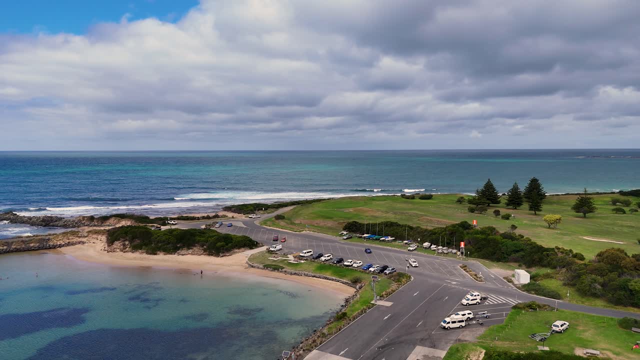 Drone footage captures Lorne's coastline, showcasing roads, ocean, and greenery under cloudy skies. The camera smoothly pans, highlighting the serene landscape
