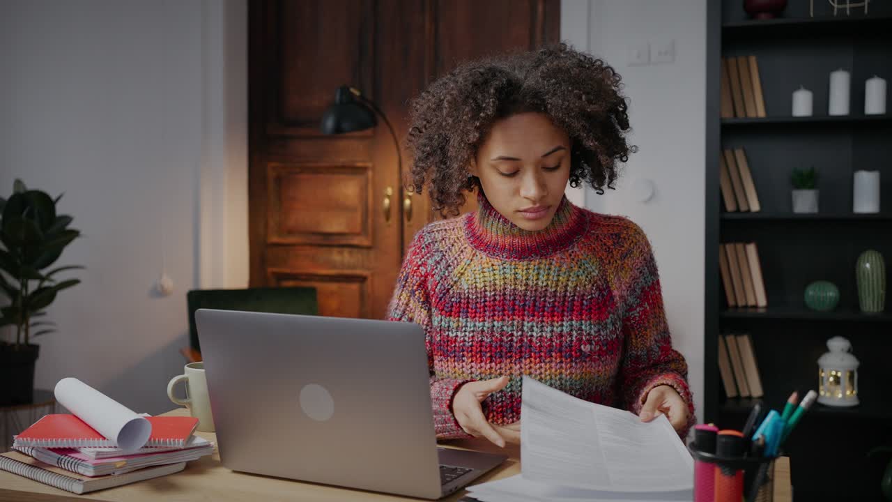 Young curly african american lady freelancer reading documents and working on laptop from home, tracking shot