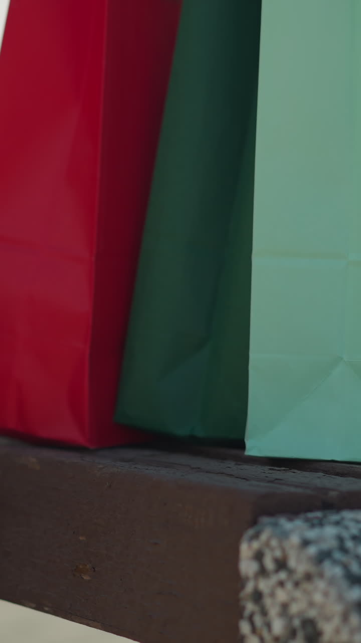 vista de la pierna de un individuo dejando caer bolsas de compras coloridas en un banco mientras está sentado, con una vista parcial de la mano descansando en su pierna