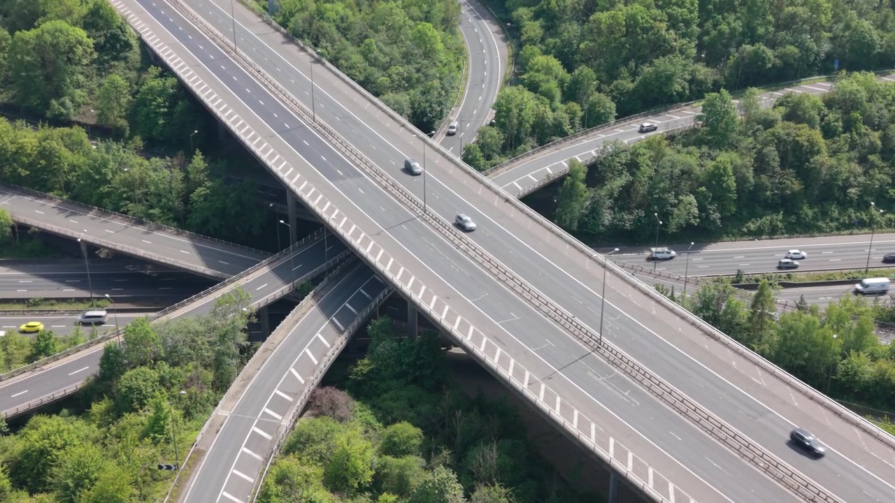 Perspectiva aérea que muestra coches circulando en un nudo de autopistas de varios niveles. Los vehículos transitan por carreteras elevadas y rampas de conexión, rodeados de abundantes árboles verdes durante el día