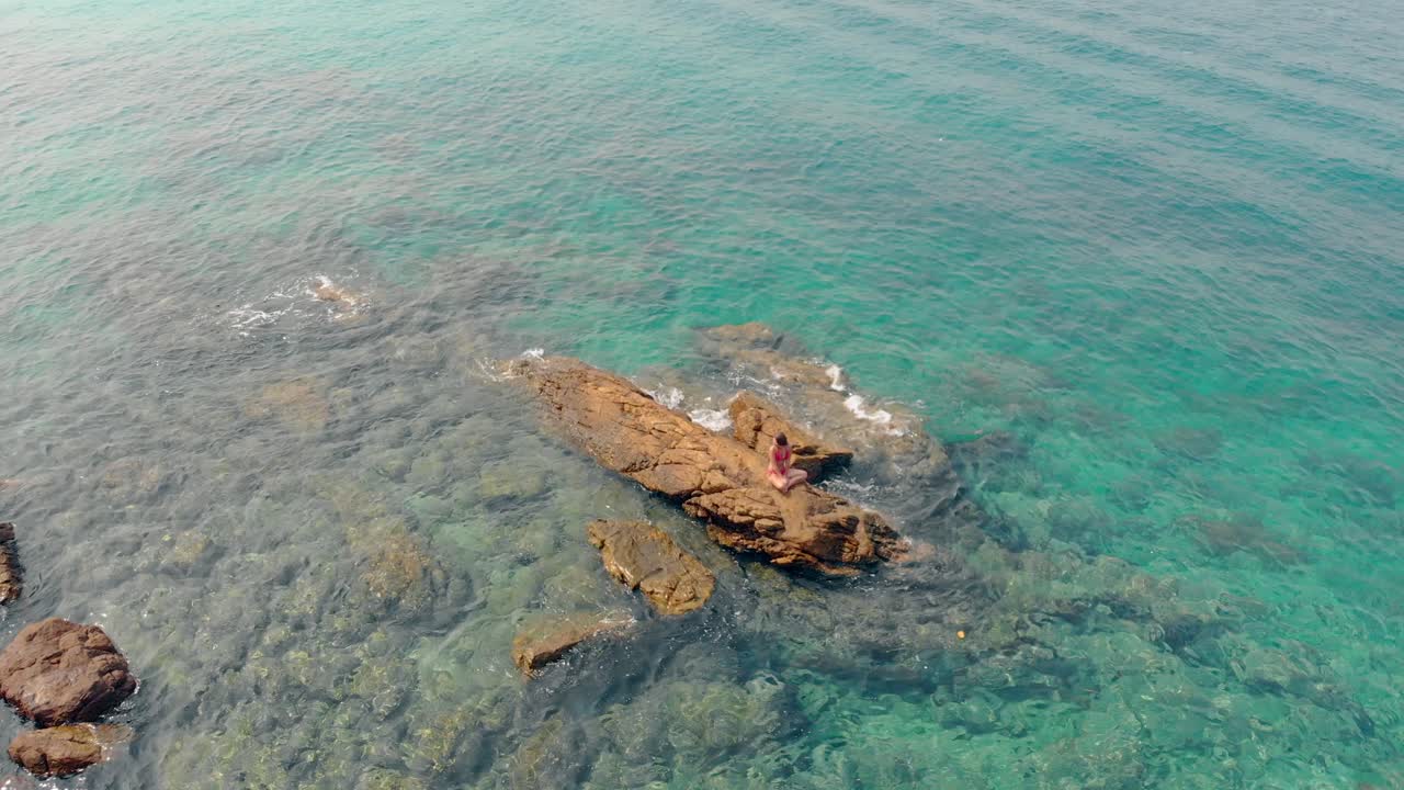 una ragazza in bikini rosso si siede su un'enorme pietra in mare agitando la mano