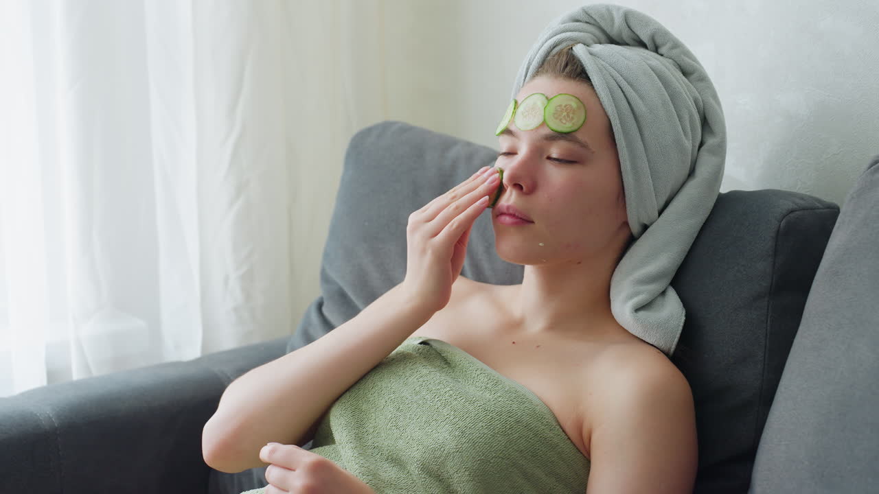 Young woman relaxing on couch wrapped in towel applying cucumber slices on face as part of skincare routine, beauty therapy and self-care at home for healthy, glowing skin
