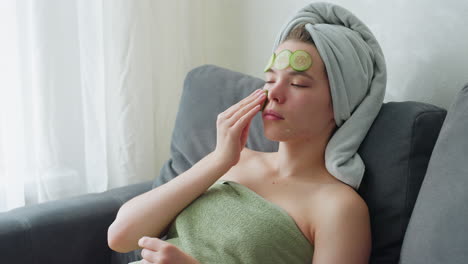 young woman relaxing on couch wrapped in towel applying cucumber slices on face as part of skincare routine, beauty therapy and self-care at home for healthy, glowing skin