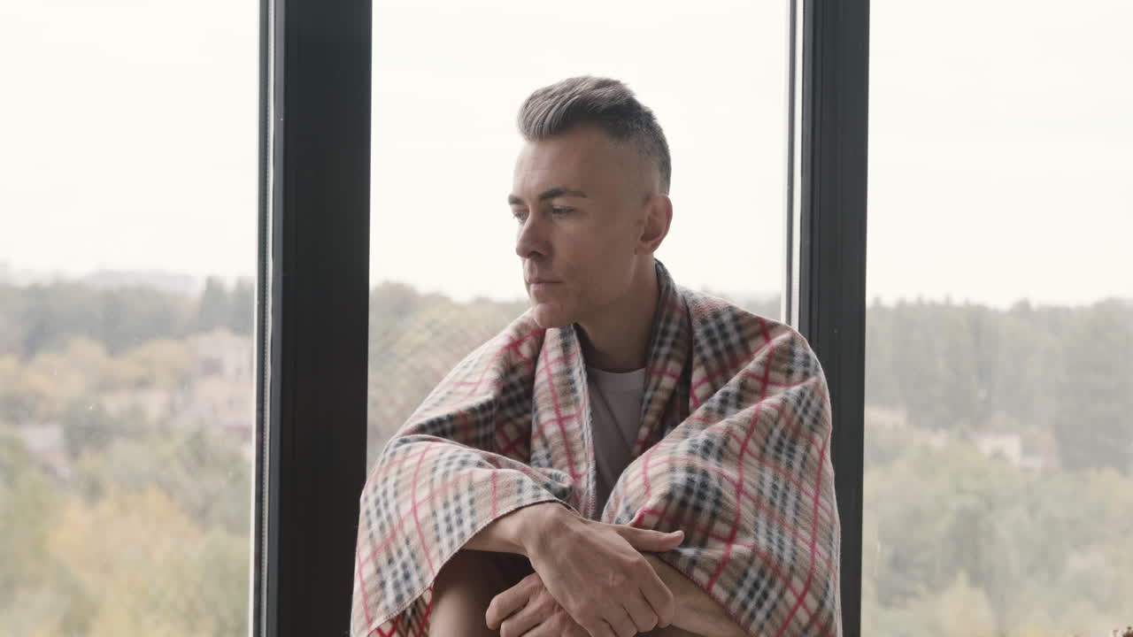 Sick Middle Aged Man With Blanket On His Shoulder Sitting At Window While His Woman Giving Him A Hot Drink And Taking Care Of Him