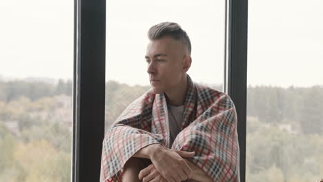 Sick-Middle-Aged-Man-With-Blanket-On-His-Shoulder-Sitting-At-Window-While-His-Woman-Giving-Him-A-Hot-Drink-And-Taking-Care-Of-Him