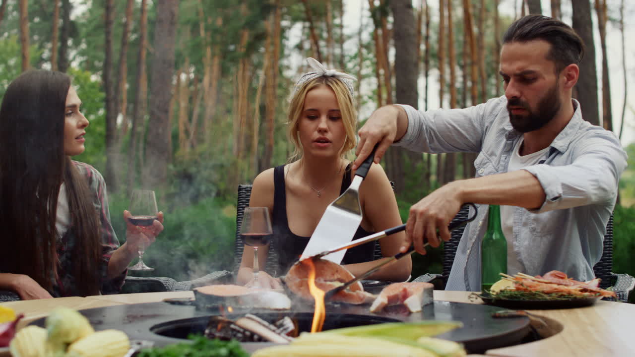 un tipo serio haciendo comida en la parrilla para una fiesta de barbacoa afuera. un tipo usando herramientas de cocina