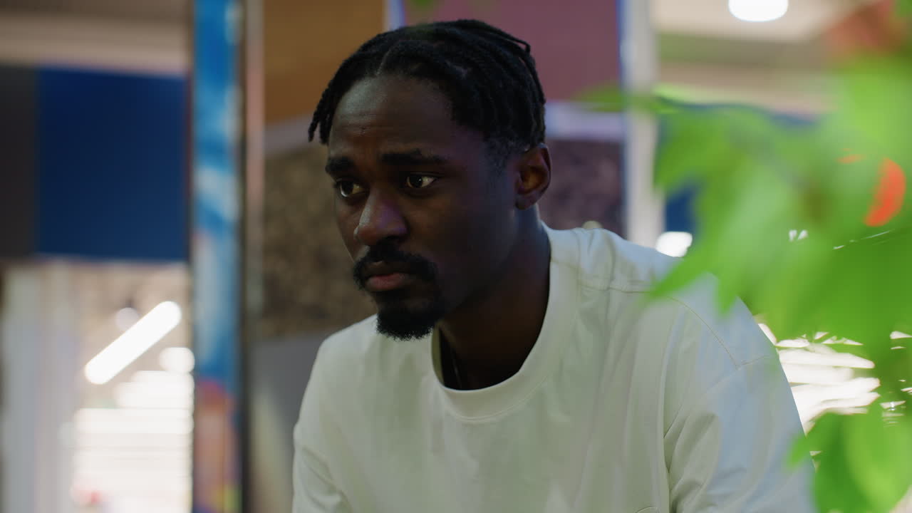 Close up of young man with braided hair wearing white shirt seated indoors in shopping mall with thoughtful expression checking time while waiting for someone near green plant