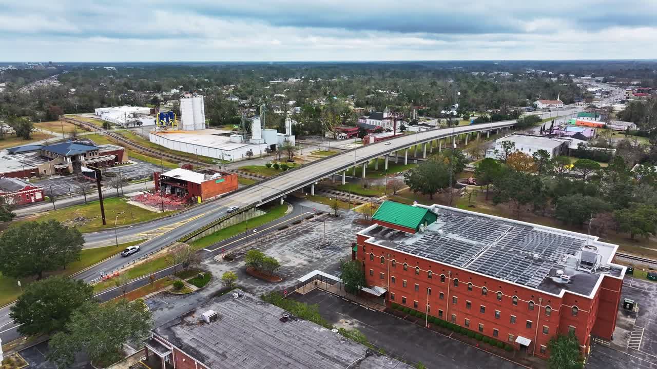 Driving cars on bridge crossing railway in Valdosta City, Georgia during cloudy day. Industrial area with factory buildings and green trees. Aerial Forward wide shot.