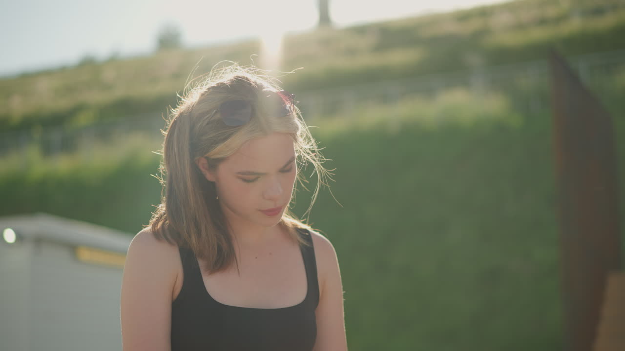 바람이 그녀의 머리카락을 불어넣는 동안 책을 읽고 앉아있는 여성, 그리고 흐릿한 배경은 언덕에 나무 구조와 레일과 함께 빛으로 목욕하는 초록색 언덕을 보여줍니다.