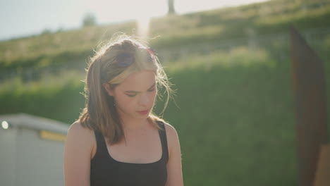 woman seated reading a book as wind blows her hair, and the blurred background shows a green hill bathed in sunlight with wooden structure and rail on the hill