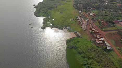 Eine-Luftaufnahme-Von-Oben,-Die-Auf-Ein-Kleines-Afrikanisches-Fischerdorf-Am-Ufer-Des-Viktoriasees-Herabblickt,-Wobei-Die-Sonne-Vom-Wasser-Reflektiert-Wird