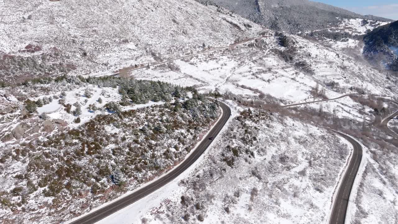 A black car drives along a winding road between snow-covered mountains, filmed with a drone