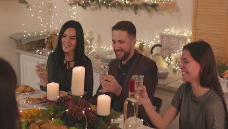 grupo de amigos felices sentados en la mesa, sosteniendo copas de champán y hablando entre ellos en casa en navidad