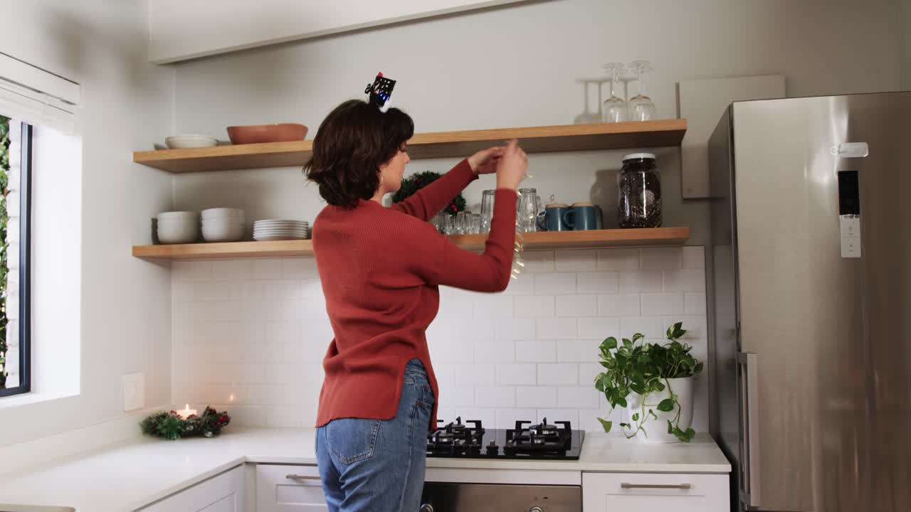 Una donna entra in cucina portando una ghirlanda, la fissa sotto una mensola e drappeggia le luci per decorare