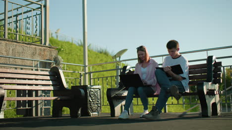 lady smiling while typing with boy focused on his tablet both seated on a bench, surrounded by greenery and light poles in the background