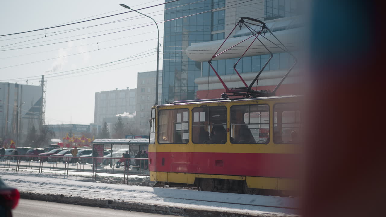 City bus at bus stop during winter commute, cars moving past on street beside modern glass building, overhead wires and urban skyline in cold daylight, passengers visible through windows