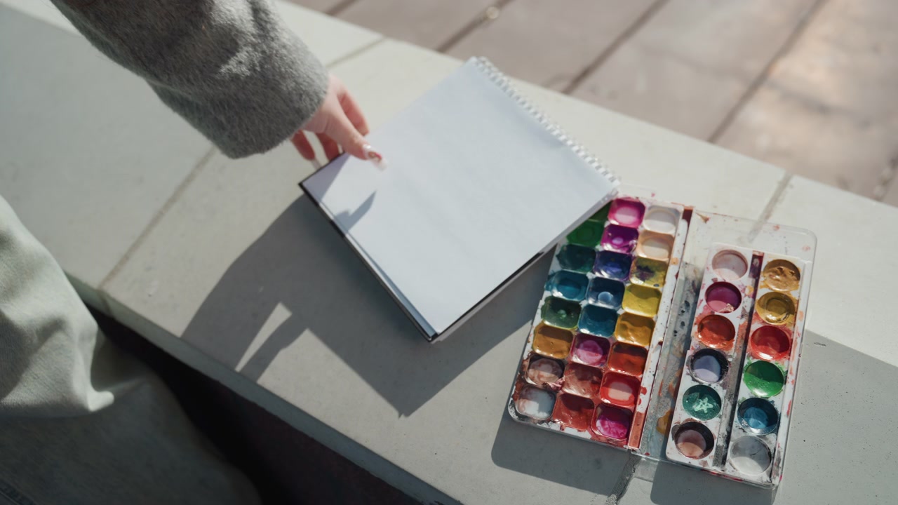 太陽の光が穏やかなスケッチシーンを照らす。カラフルな水彩絵の具セットを手に立ち止まる都会のアーティスト。鮮やかな絵の具セットとスケッチブックの傍らで休むアーティストが描かれた静かな都市公園の風景。