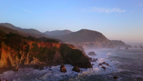 Bixby-Creek-Bridge-Big-Sur-California-aerial-cinematic-drone-flight-Pacific-Ocean-Nor-Cal-winter-summer-big-wave-swell-crashing-rugged-coastline-afternoon-golden-hour-pink-sunset-downward-motion