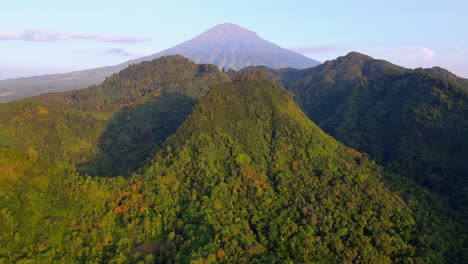 Aerial-view-of-hills-overgrown-with-rainforest-with-huge-mountain-on-the-background-in-blue-sky