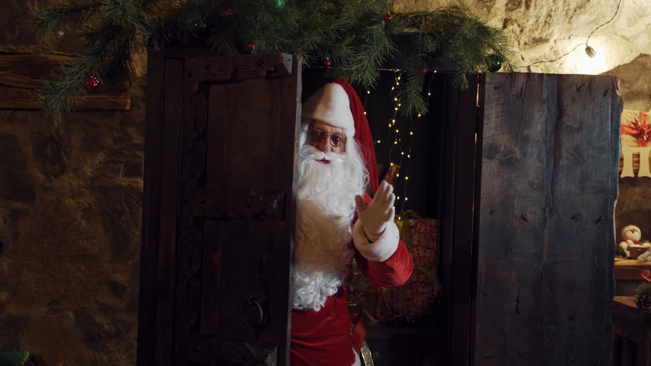 Santa look out from the door and waving hand in white glove. Wooden door and Santa Claus with white beard and eyeglasses making bye gesture. Christmas time.