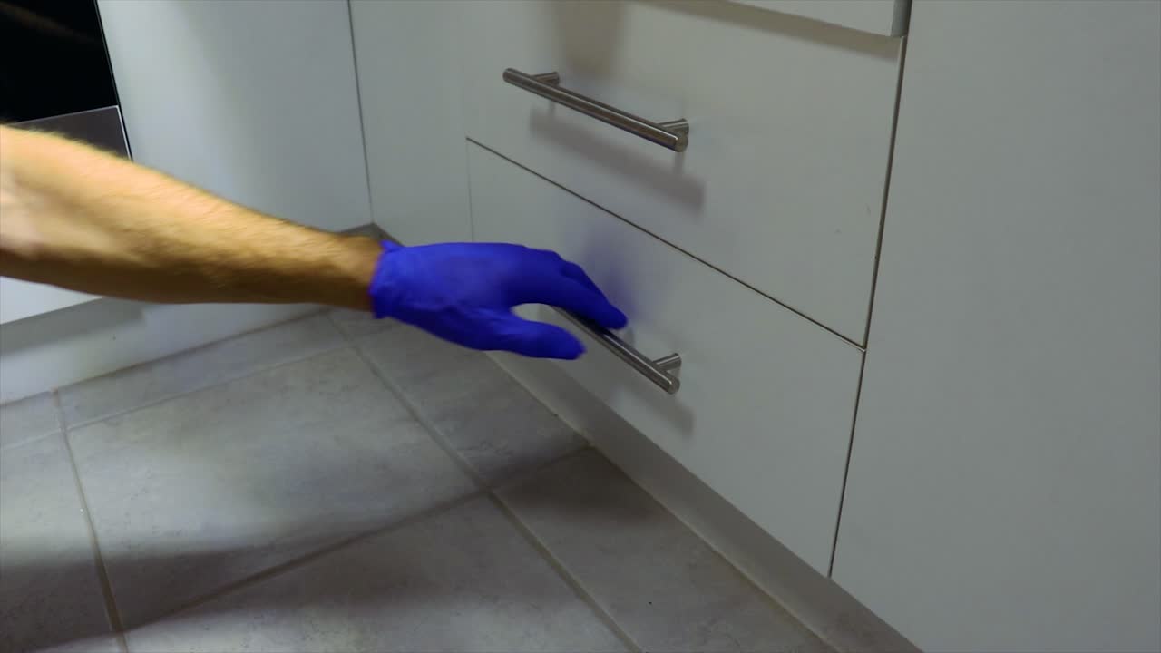 European Male with blue glove on opens white kitchen drawer with silver handle to reveal a large jagged knife with a black handle at a crime scene